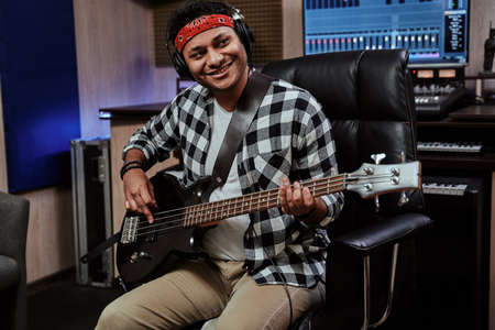 Portrait of happy young man, male artist in headphones smiling aside, playing guitar while sitting in recording studioの写真素材