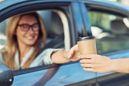 But first coffee. Happy caucasian businesswoman sitting behind steering wheel of her modern car and buying take away coffee, focus on the paper cupの写真素材