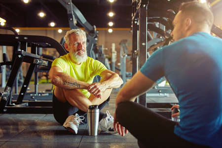 Resting after training. Athletic middle aged man discussing something with fitness instructor or personal trainer while sitting together on the floor at gymの写真素材