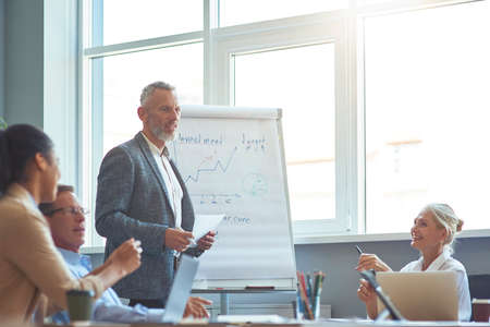 Explaining business strategy. Confident mature businessman standing near flipchart, giving a presentation while having a brainsportm meeting with team in the officeの写真素材