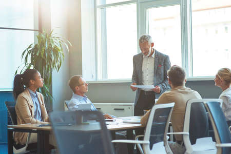 Confident mature businessman in formal wear holding document and discussing project results with multicultural team, having a meeting in the modern officeの写真素材