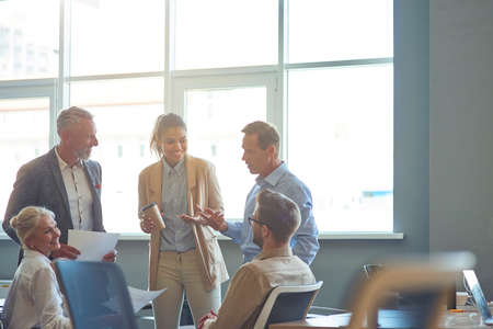 Powerfully successful team. Group of multiracial business people in classic wear discussing work and sharing ideas while standing in the modern office or coorking spaceの写真素材