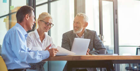 Successful business people working together, three happy coworkers interacting, discussing something and smiling while sitting at desk in the modern office or coworking spaceの写真素材