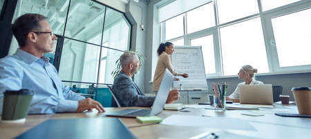 Young mixed race woman writing something on flipchart, giving a presentation and discussing business strategy with colleagues, startup team working together in the modern officeの写真素材