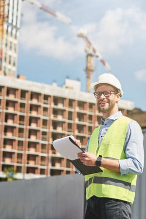 Good job. Young happy civil engineer or construction supervisor wearing helmet looking away and smiling while inspecting building site and making some notesの写真素材