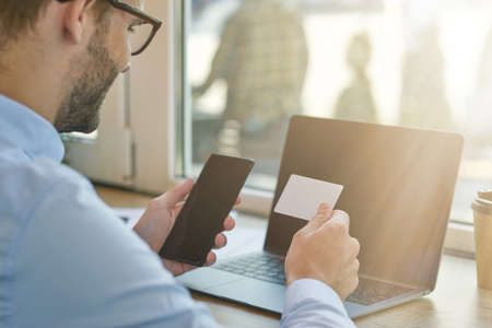 Important call. Rear view of a young businessman or male freelancer holding smartphone and looking at blank business card while working remotely in cafeの写真素材