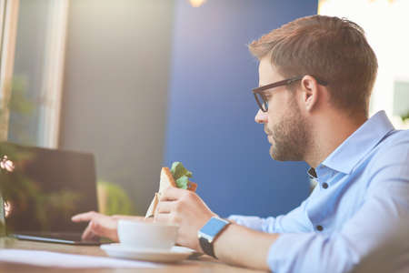Side view of a young businessman sitting at wooden table in cafe, using laptop and eating fresh sandwichの写真素材