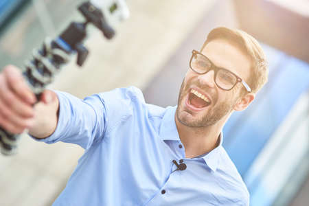 Creating new content. Young happy male blogger wearing blue shirt and eyeglasses holding a gimbal with smartphone and recording video for his blog while standing outdoorsの写真素材
