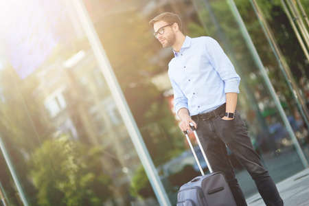 Business travel. Elegant handsome caucasian businessman with suitcase looking away while waiting taxi outdoorsの写真素材