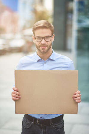 Vertical shot of a young serious man, male activist wearing blue shirt and eyeglasses holding empty sign board and looking at camera while protesting outdoorsの写真素材