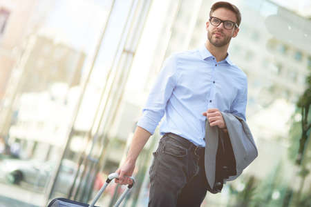 Young confident businessman carrying his luggage and looking away while standing outdoors, arriving at airportの写真素材