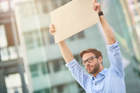 Protesting outdoors. Portrait of a young male activist wearing blue shirt and eyeglasses holding empty sign board over the head while standing outdoorsの写真素材