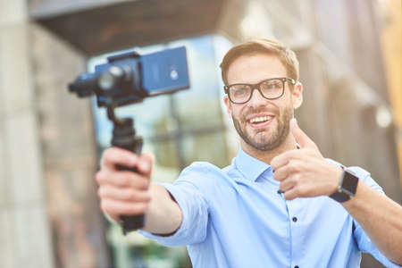 Young cheerful male blogger wearing blue shirt and eyeglasses holding a gimbal with smartphone, smiling and showing thumb up while recording new video for his blog outdoorsの写真素材