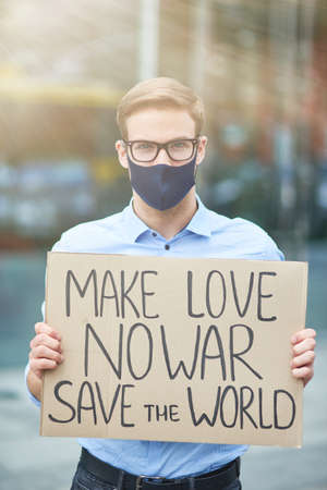 Vertical of young male activist wearing black protective mask holding banner or poster and looking at camera while protesting against the war outdoors, standing on the roadの写真素材