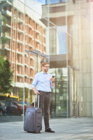 Vertical shot of elegant handsome businessman with suitcase using smartphone and looking away while standing outdoors, waiting for taxiの写真素材