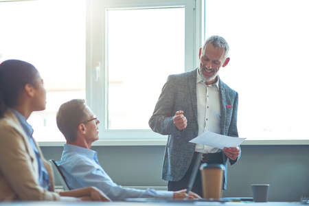 Confident mature male executive in formal wear holding document and discussing project results with young colleagues. Business people working together in the modern officeの写真素材