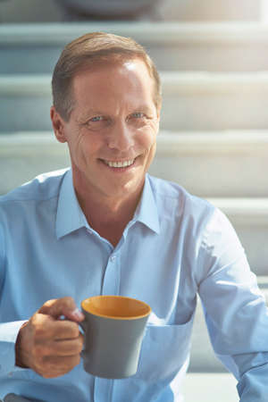 Happy mature businessman in shirt holding cup of coffee, looking at camera and smiling while having a break in the office, vertical shotの写真素材