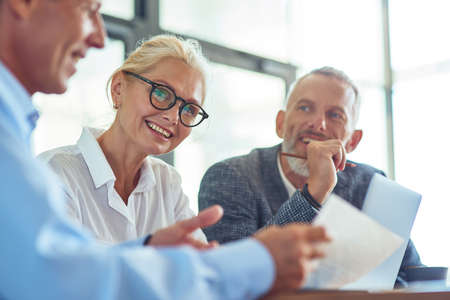Beautiful mature businesswoman wearing eyeglasses looking at camera and smiling while discussing contract with partners in the modern officeの写真素材