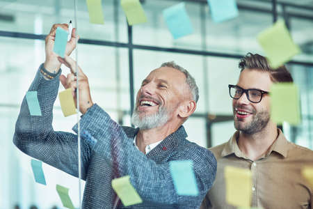 Two happy male coworkers putting colorful sticky notes on a glass wall and smiling while standing in the modern officeの写真素材