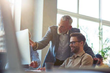 Business people sitting at desk in the modern office, looking at PC screen and discussing project, confident mature man explaining something to his young male coworkerの写真素材