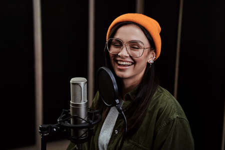 Portrait of cheerful young woman, hip hop artist singing into a condenser microphone while recording a song in a professional studioの写真素材