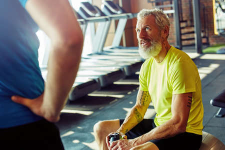 Feeling exhausted. Caucasian middle aged man in sportswear holding bottle of water and talking with personal trainer while resting after workout at gym or sport clubの写真素材