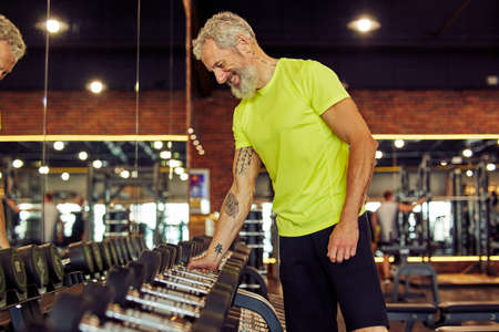 Side view of a handsome mature man in sportswear taking a dumbbells while working out at gym, exercising with weightsの写真素材