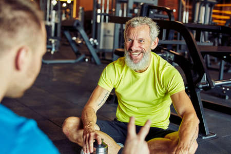 Happy middle aged man discussing something with fitness instructor or personal trainer and smiling while sitting together on the floor at gymの写真素材