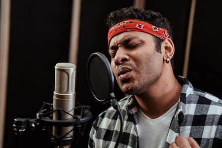 Portrait of young man, hip hop artist looking focused, singing into a condenser microphone while recording a song in a professional studioの写真素材