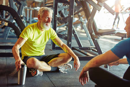 Talking with personal trainer. Happy middle aged man discussing something with fitness instructor and smiling while sitting together on the floor at gymの写真素材