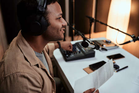 Portrait of young male radio host going live on air, talking in microphone, holding a script paper while sitting in studioの写真素材