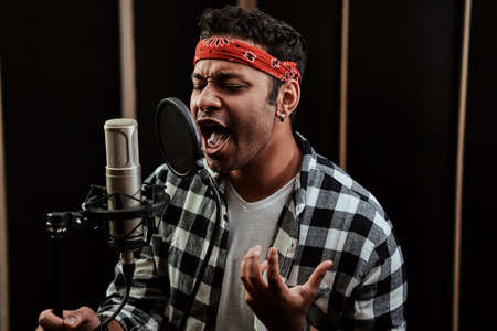 Portrait of young man, hip hop artist singing with closed eyes into a condenser microphone while recording a song in a professional studioの写真素材