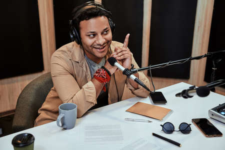 Portrait of happy young male radio host smiling while talking, broadcasting in studio using microphone and headphonesの写真素材