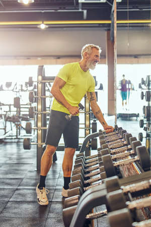 Full length shot of a happy athletic mature man in sportswear taking a dumbbells, looking in a big mirror and smiling while exercising with weights at gymの写真素材