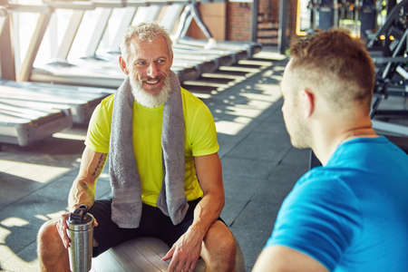 Talking a break. Positive middle aged man in sportswear holding bottle of water and talking with fitness instructor at gym or sport clubの写真素材