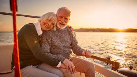 Enjoying amazing sunset. Happy senior couple, elderly man and woman holding hands, hugging and relaxing together while sitting on the side of yacht deck floating in the seaの写真素材