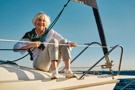 Enjoying sea trip. Happy senior woman sitting on the side of a sail boat or yacht deck floating in sea, looking at camera and smiling. Clear blue sky backgroundの写真素材