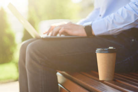 Cropped shot of a male freelancer working online on laptop and drinking coffee while sitting on the bench outdoors on a sunny day, side viewの写真素材