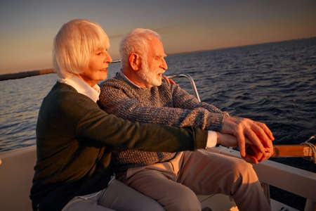 Side view of beautiful senior couple sailing at sunset, elderly man and woman sitting on the side of boat or yacht deck floating in the seaの写真素材
