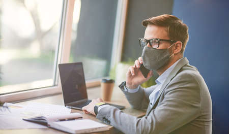 Side view of a busy businessman wearing protective face mask using laptop and talking by phone with partner while working remotely in cafe during covid 19 outbreakの写真素材