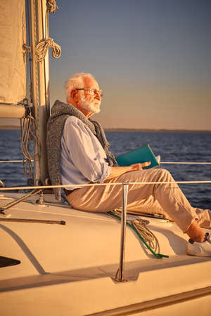 Vertical shot of relaxed retired man reading book and enjoying amazing sunset while sitting on the side of his sailboat or yacht deck floating in the calm blue seaの写真素材