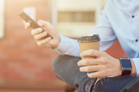Cropped shot of a businessman in blue shirt using smartphone and drinking coffee while relaxing on the bench outdoorsの写真素材