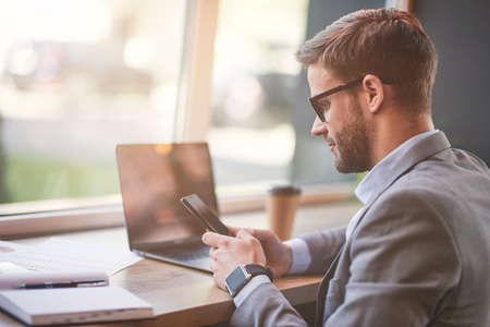 Side view of a young businessman or male freelancer using his smartphone while working online remotely in cafeの写真素材