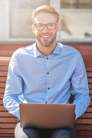 Vertical shot of a young happy businessman using laptop, looking at camera and smiling while sitting on the bench outdoors and working onlineの写真素材