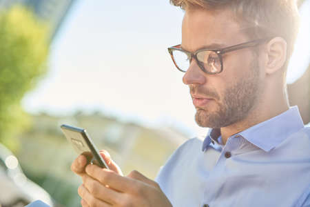 Side view of a young handsome businessman using smartphone while sitting on the bench outdoors on a sunny dayの写真素材