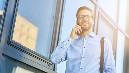 Good news. Young happy caucasian businessman in blue shirt talking by phone and smiling while standing outdoors. Business people and communication conceptの写真素材