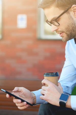 Taking a break at work. Side view of a handsome businessman using smartphone and drinking coffee while relaxing on the bench outdoorsの写真素材