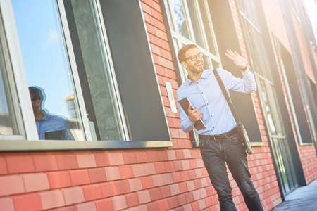 Young happy caucasian businessman wearing blue shirt and eyeglasses holding his laptop and waving, saying hello while standing against office building outdoorsの写真素材