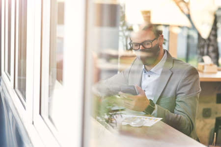 Work during covid 19 outbreak. Young businessman or male freelancer wearing protective face mask using smartphone while working remotely in cafeの写真素材