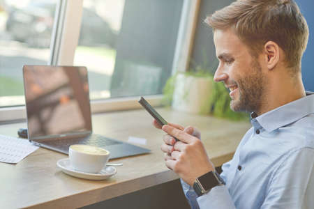 Chatting with friends. Side view of a freelancer or businessman using his smartphone and smiling while working remotely in cafe, drinking coffeeの写真素材